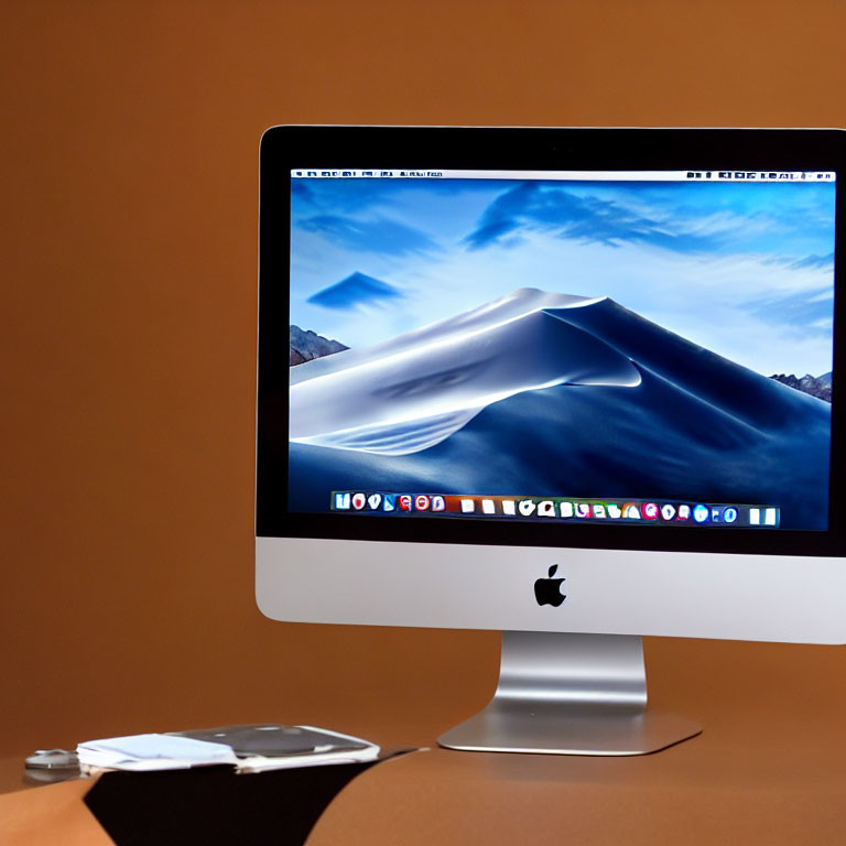 Desktop setup with iMac, desert dune wallpaper, smartphone, and glasses on brown desk