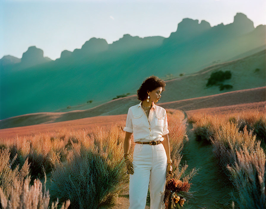 Woman in white outfit walking through desert with purple foliage and mountains in background
