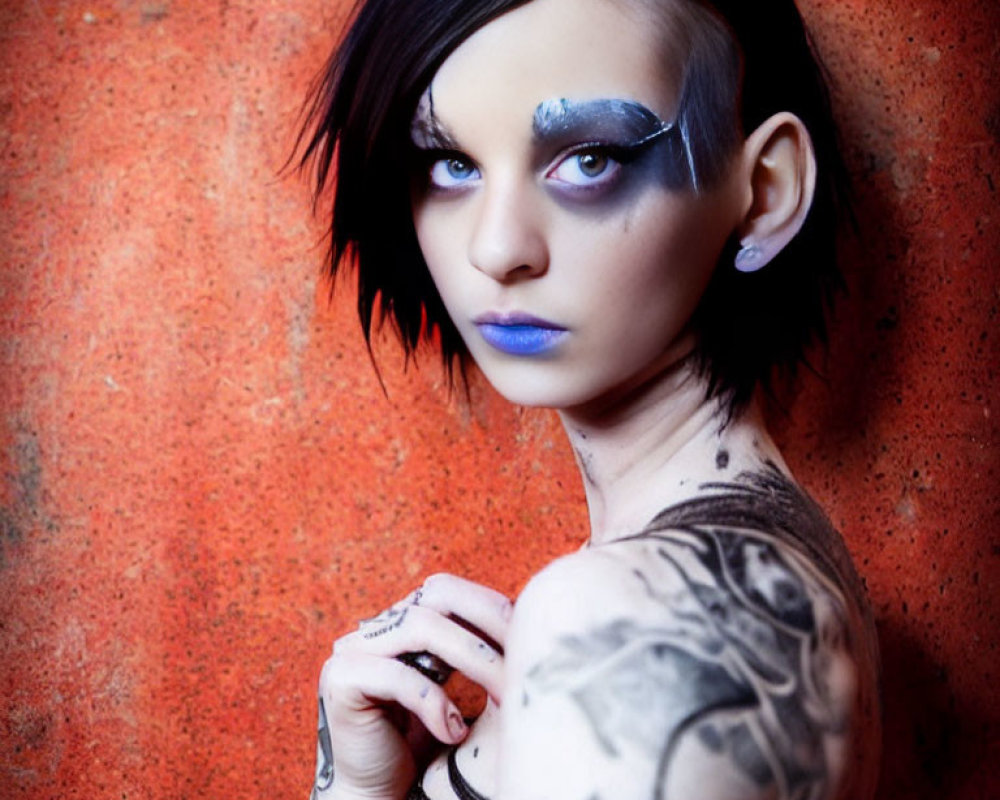 Portrait of person with dark hair, silver eye makeup, blue lipstick, and tattoos on orange background