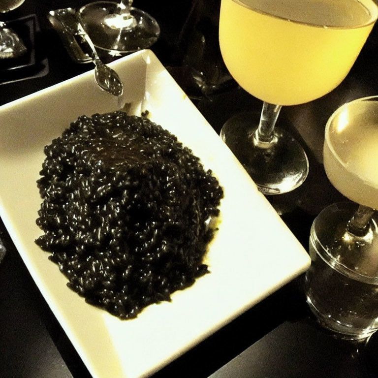 Golden-hued black risotto plate with yellow beverages on dark table setting