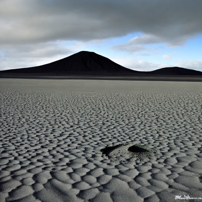 Desolate landscape with rippled sand and volcanic cone under cloudy sky