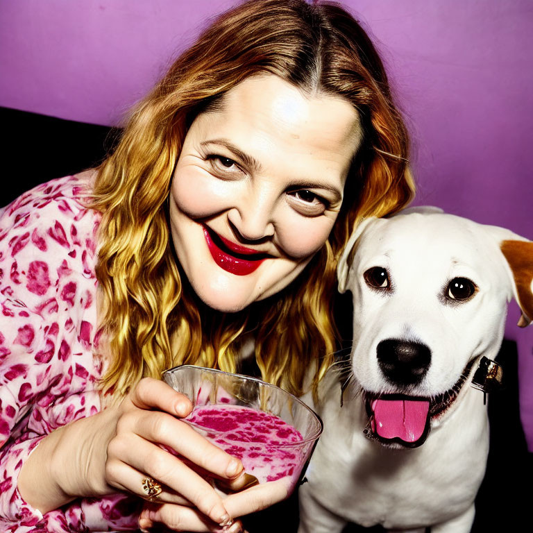Smiling woman with red lipstick and wavy hair holding pink drink next to happy white dog