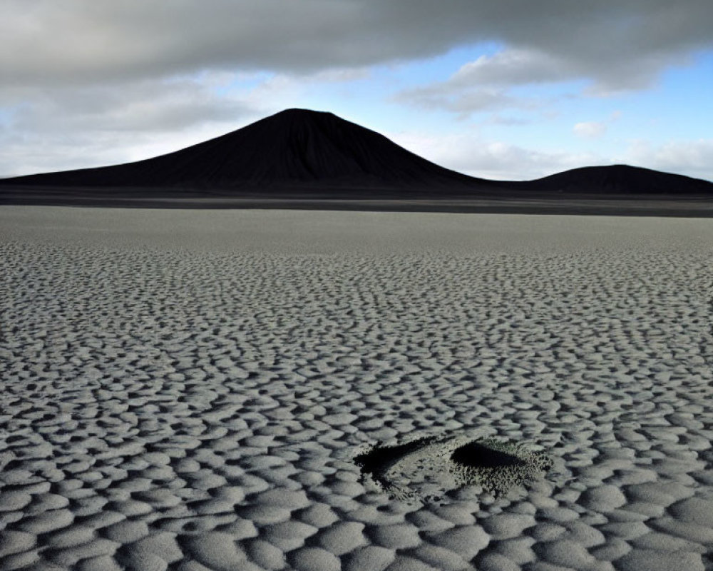 Desolate landscape with rippled sand and volcanic cone under cloudy sky