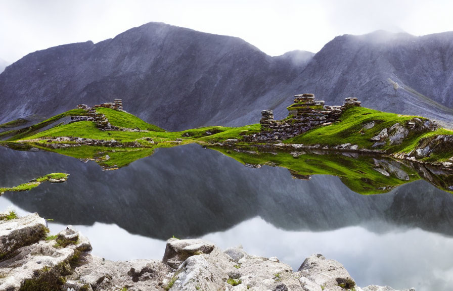 Ruined stone structures on lush green hillside with clear reflection in mountain lake surrounded by foggy peaks