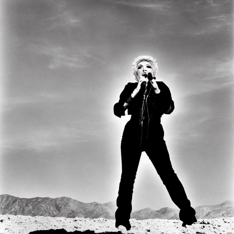 Person posing with microphone in barren landscape under dramatic sky