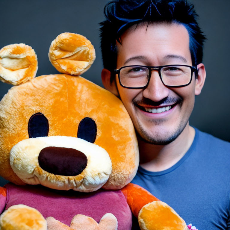 Smiling person with glasses holding large teddy bear on grey backdrop