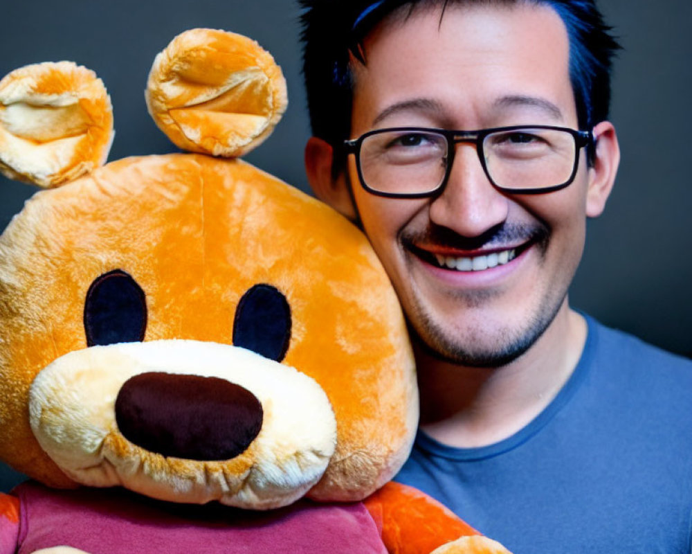 Smiling person with glasses holding large teddy bear on grey backdrop