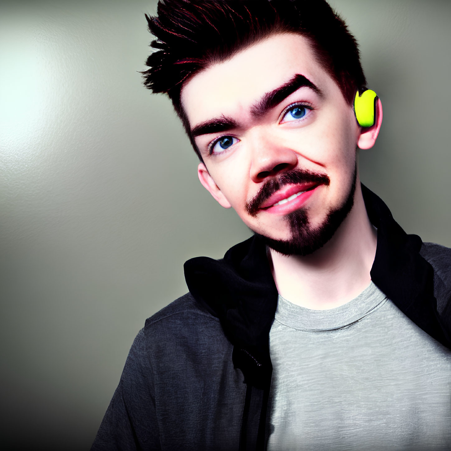 Young adult with styled hair, goatee, earring, grey shirt, and black hoodie smiling at