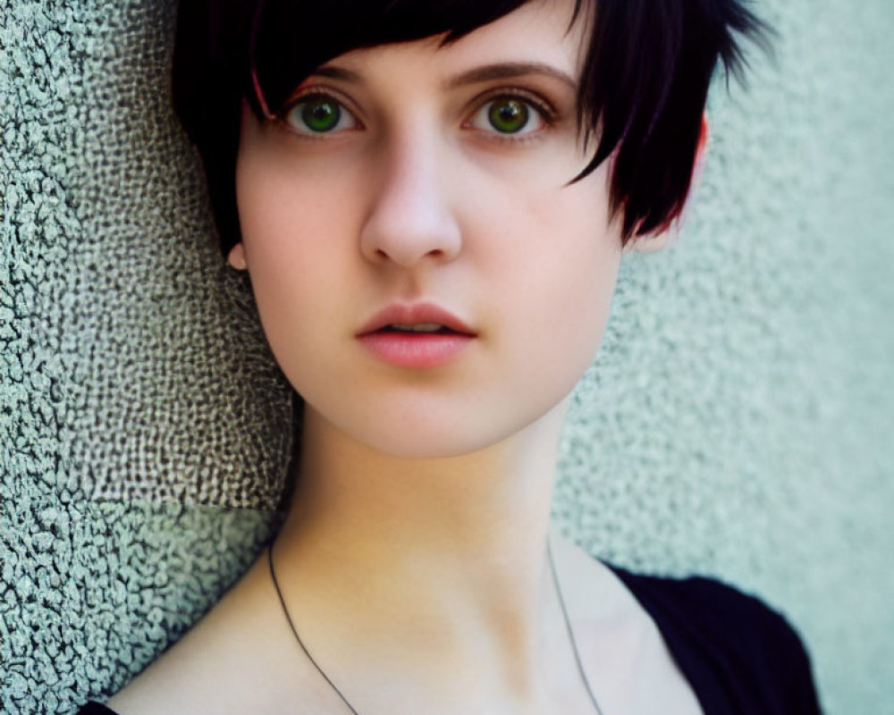 Black-haired person with green eyes in black top against textured wall.