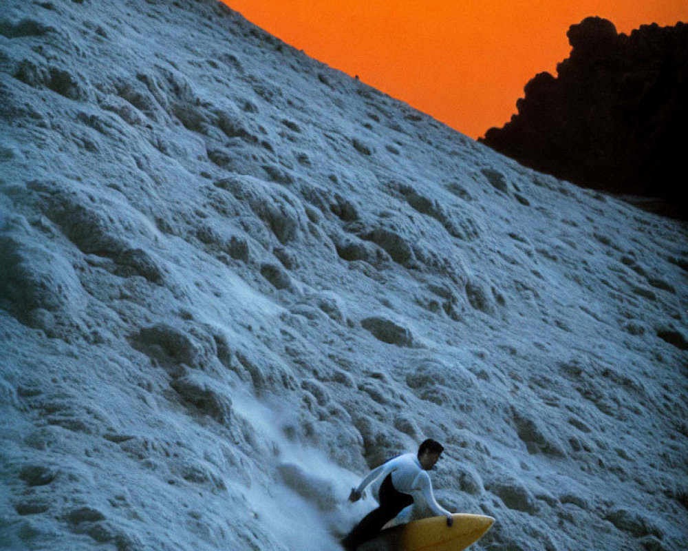 Surfer riding large wave against orange sky with foamy waters and rocks in background