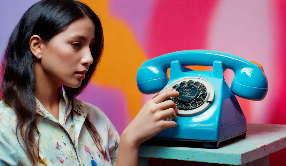 Woman in Paint-Splattered Shirt Dialing Retro Blue Rotary Phone on Vibrant Background