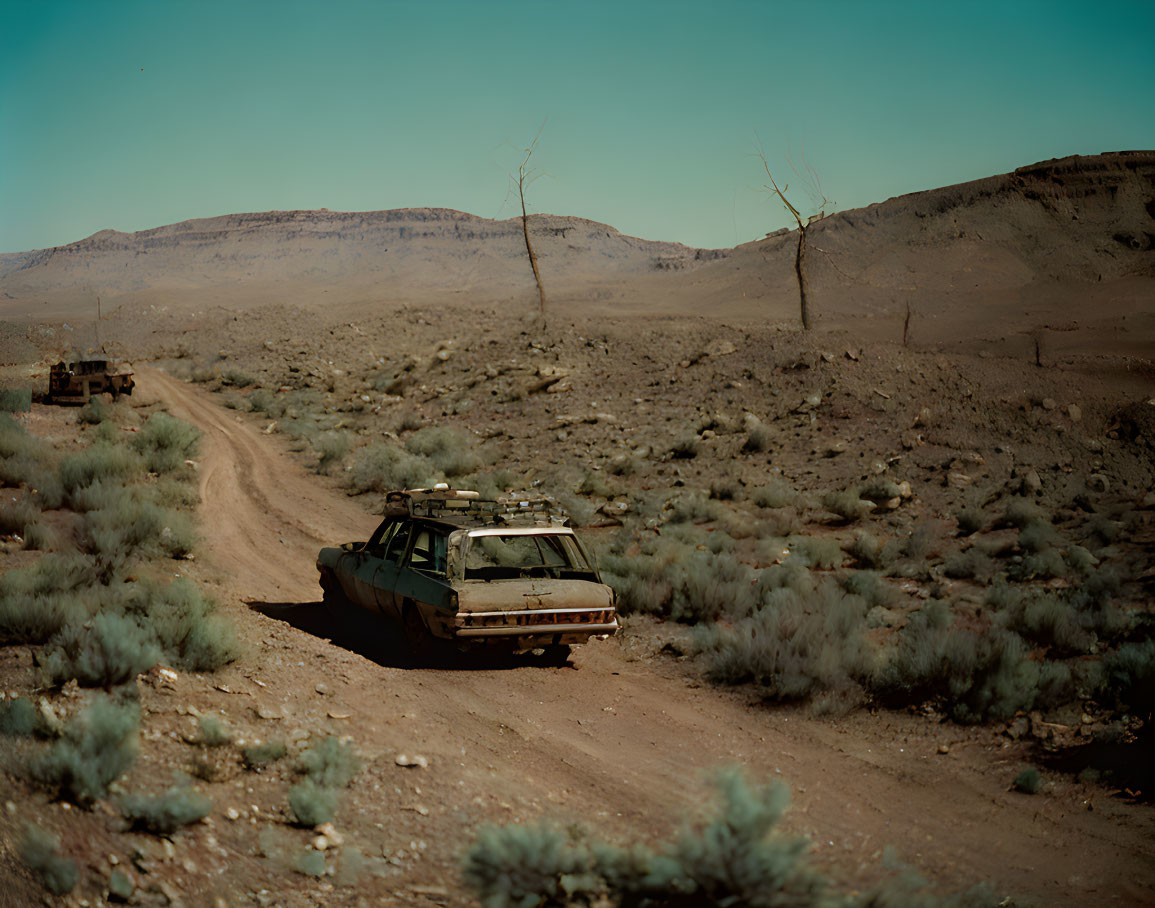 Abandoned car on dusty trail with arid hills and sparse vegetation.