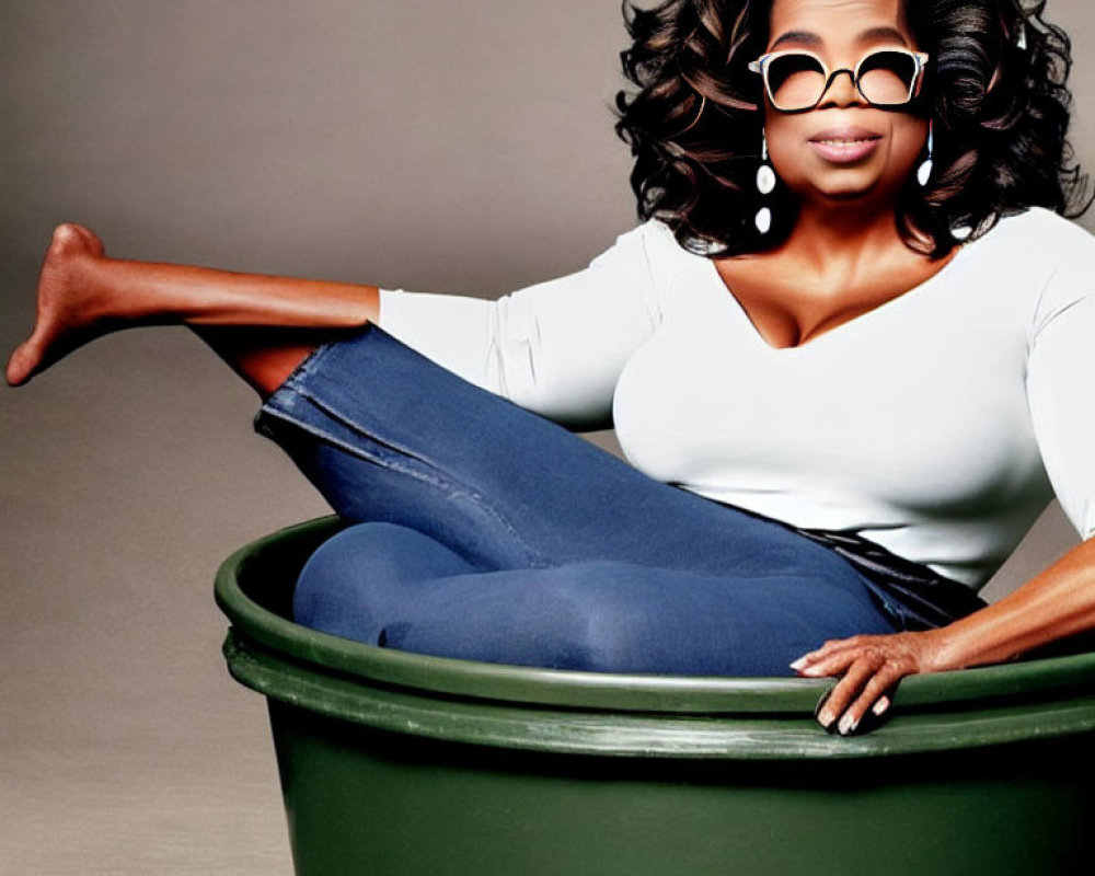 Person in sunglasses sits inside green trash can with white top and blue jeans