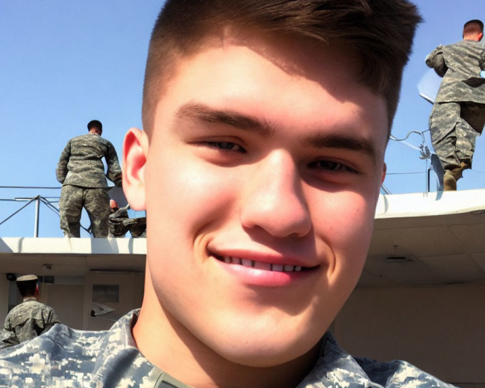 Young man in military uniform smiling with others in background under clear skies