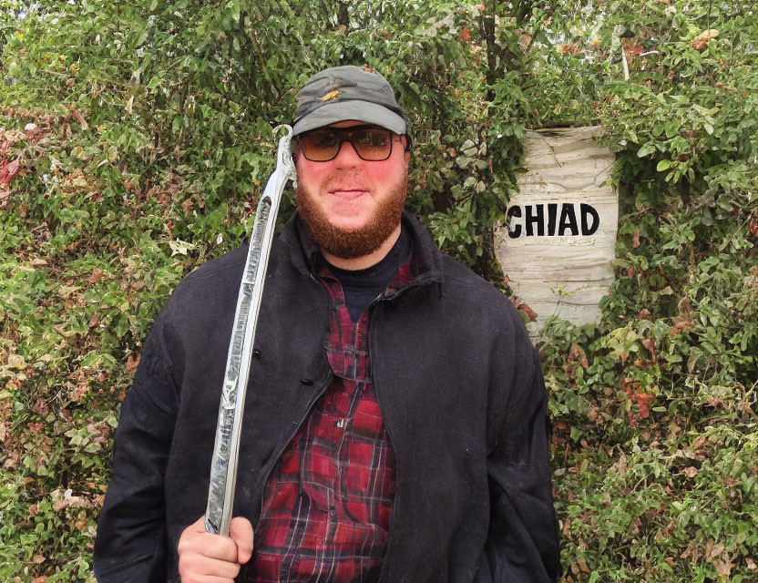 Bearded man in sunglasses and cap with "CHIAD" sign in front of dense foliage