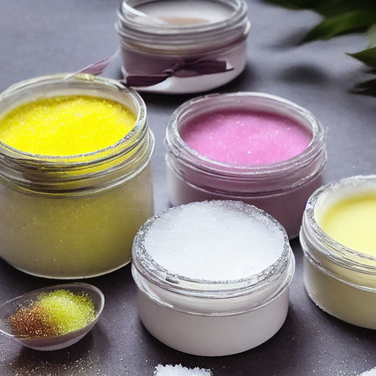 Colorful Lip Scrubs in Open Jars on Gray Surface with Granules and Green Leaves
