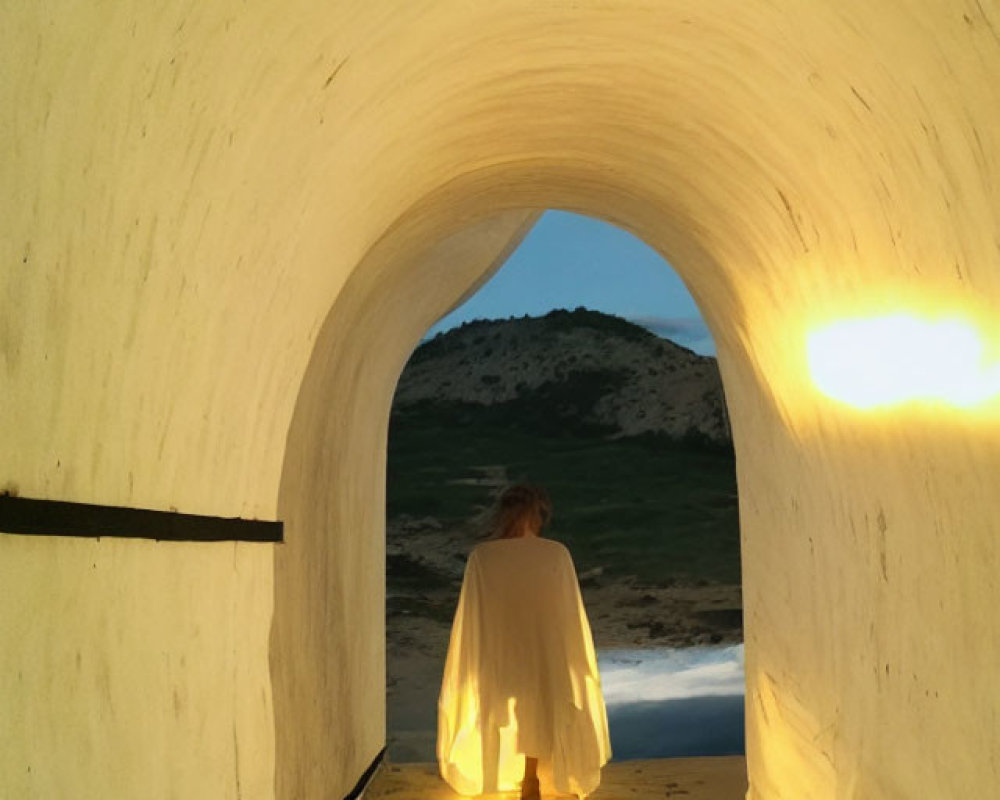Figure in white cloak gazes at landscape from tunnel's end at dusk or dawn