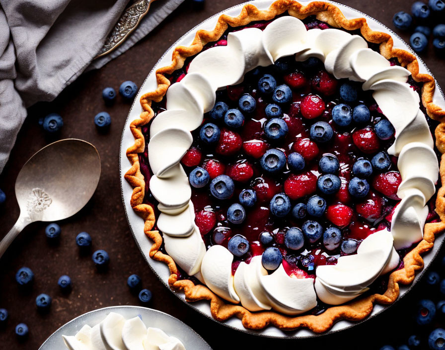 Berry tart with blueberries and raspberries, whipped cream, spoons, and loose berries on dark