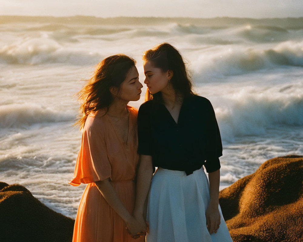 Two women in elegant attire standing close together on a beach with crashing waves.