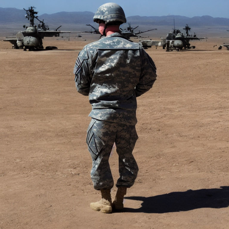 Soldier in camouflage uniform observing helicopters and military vehicles in desert setting
