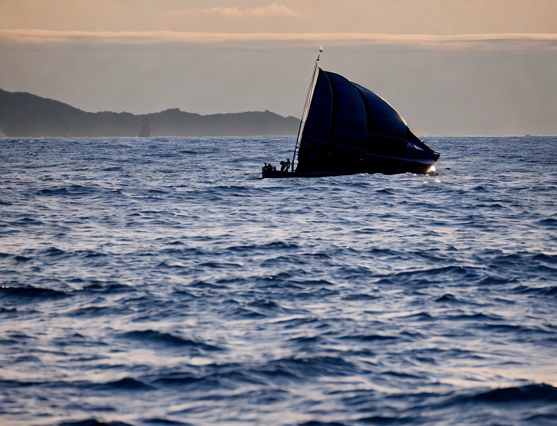Silhouetted sailboat with large blue sail on dusky ocean with land in distance