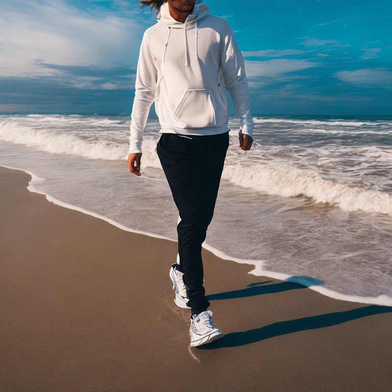 Person in White Hoodie Walks on Sandy Beach with Blue Sky and Waves
