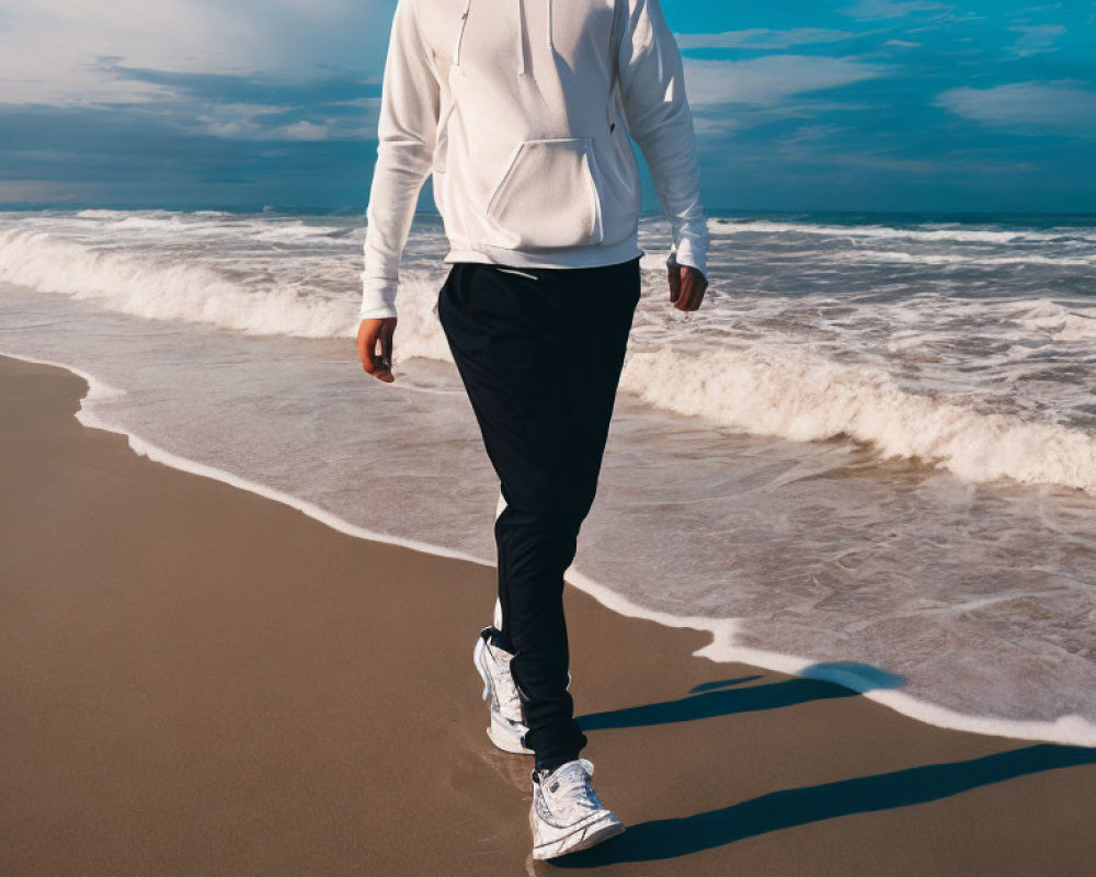Person in White Hoodie Walks on Sandy Beach with Blue Sky and Waves