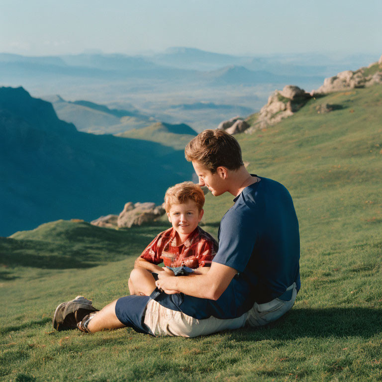 Man and boy on grassy hillside with vast mountain landscape.