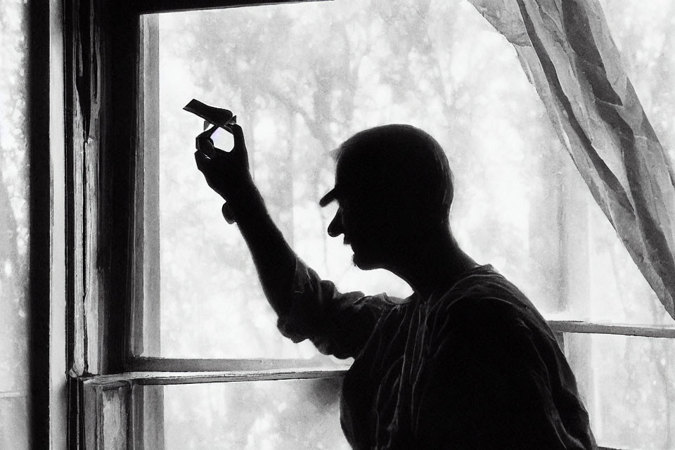 Silhouetted person holding small object by window with billowing curtains