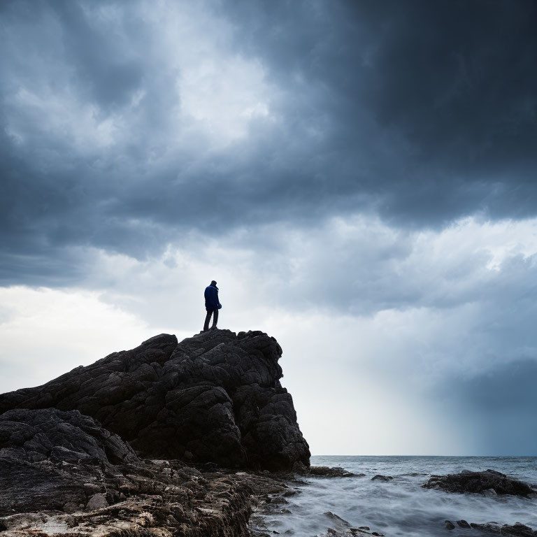 Solitary figure on rocky outcrop gazes at stormy sea.
