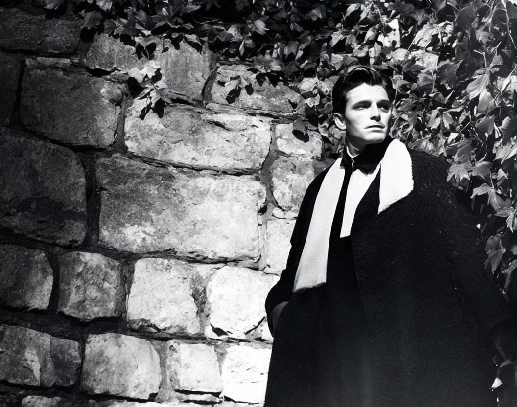 Vintage-clad man in cape against vine-covered wall in black and white.