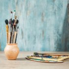 Assorted paintbrushes in golden cup on blue wall with plant art