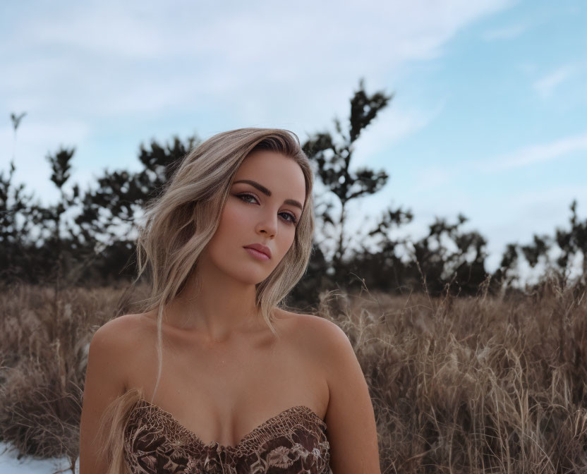 Blonde woman in lace dress standing in dry grass under cloudy sky