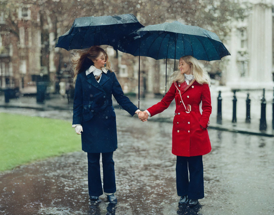 Two women holding hands under umbrellas in snowy street, one in red coat, the other in blue