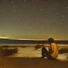 Person sitting on hill under starry night sky with town lights, comet, and moon.