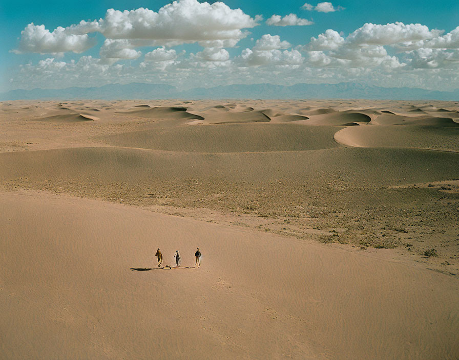 Three individuals walking in vast desert with rolling sand dunes under blue sky.
