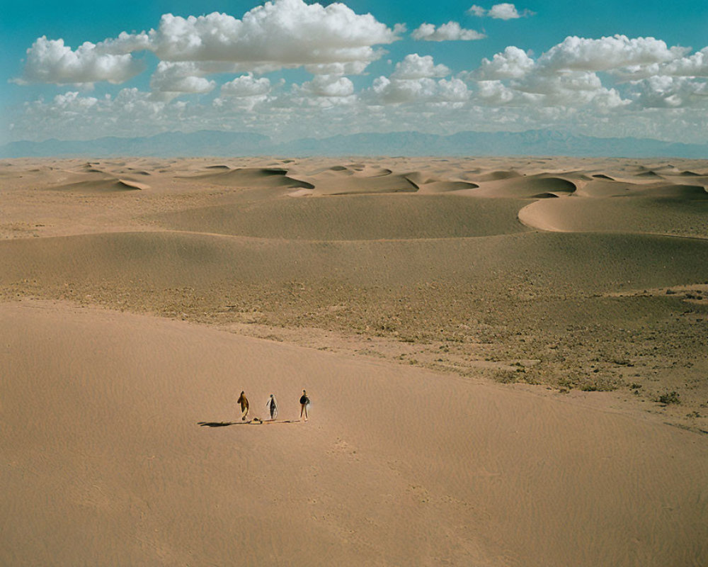 Three individuals walking in vast desert with rolling sand dunes under blue sky.