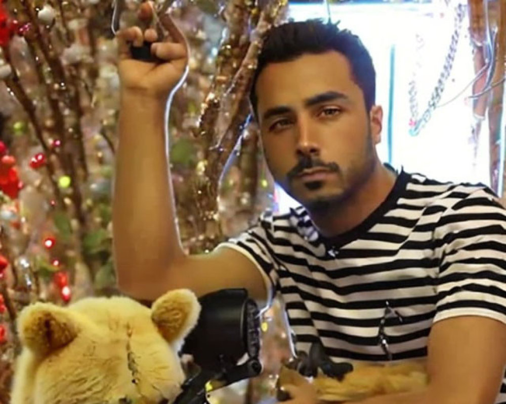 Bearded man in striped shirt with camera and prop rifle next to stuffed toy bear under twinkling lights