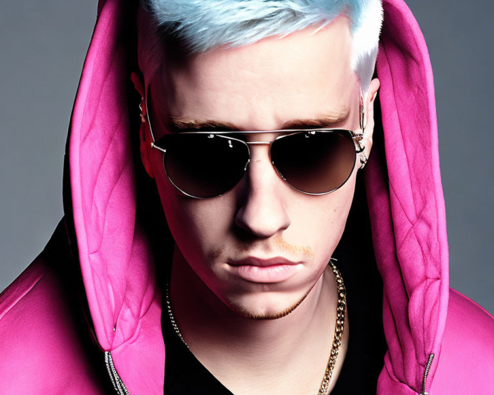 Blue-haired person in pink hoodie with aviator sunglasses and light mustache.