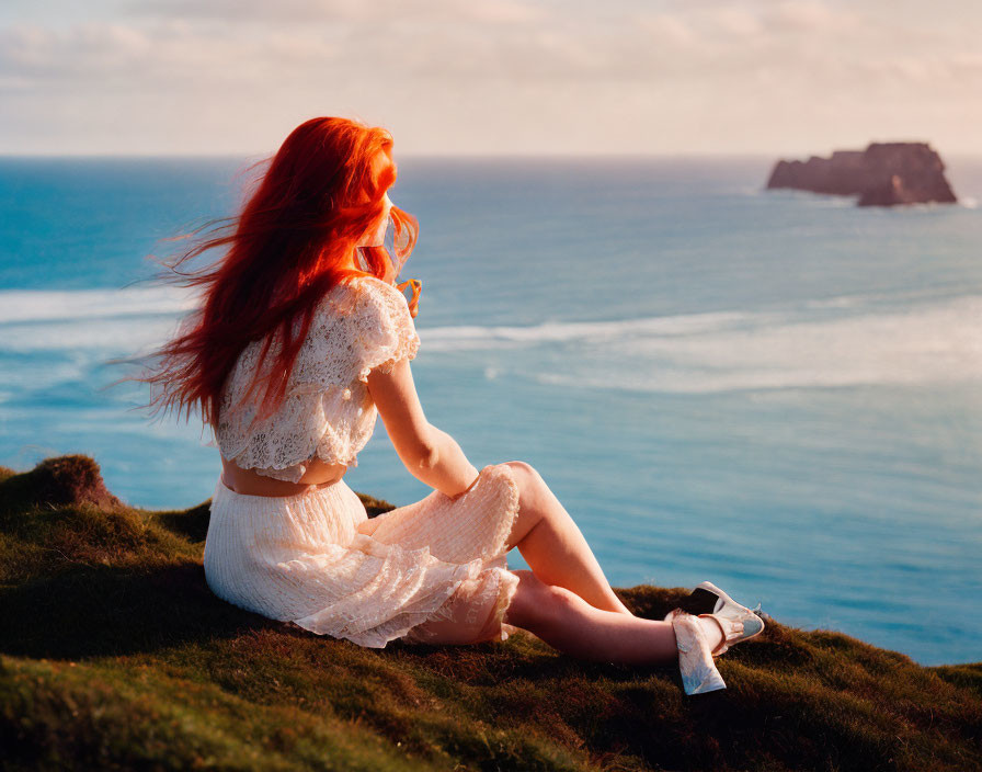 Red-haired woman in white dress gazes at sea from cliff at sunset