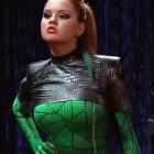 Red-haired woman in green galaxy bodysuit on dark backdrop.
