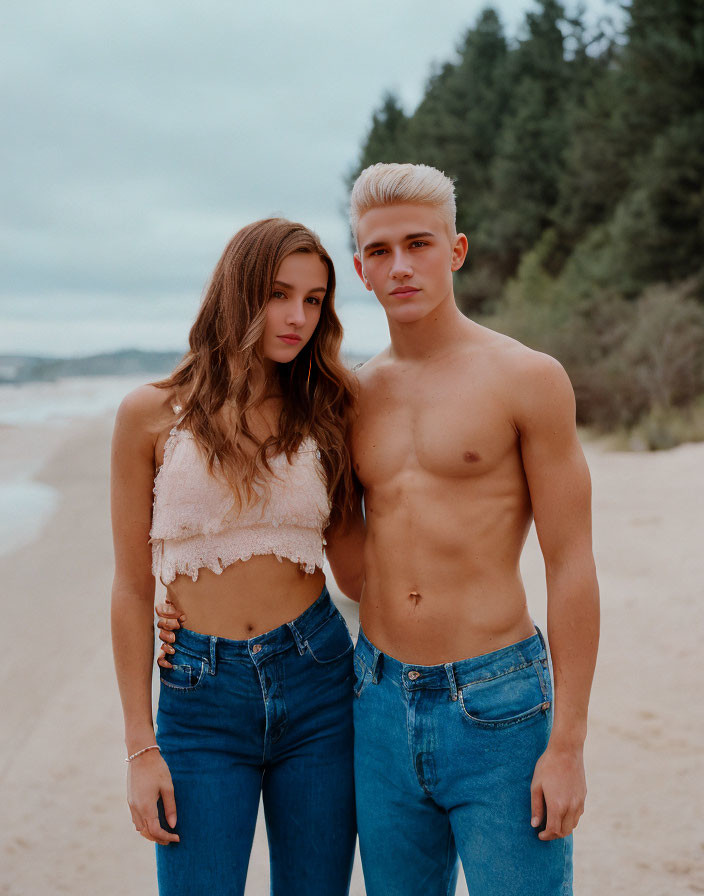 Young man and woman with platinum blonde hair on sandy beach in blue jeans and crop top.