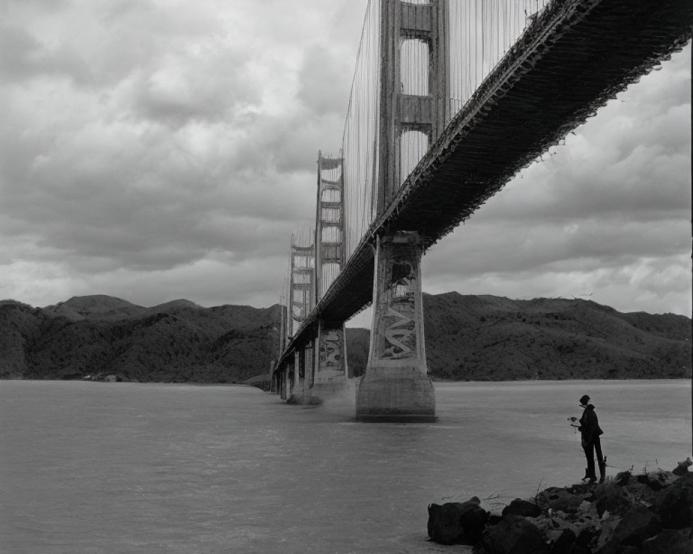 Person standing by water under suspension bridge with pillars, hills, and cloudy sky.
