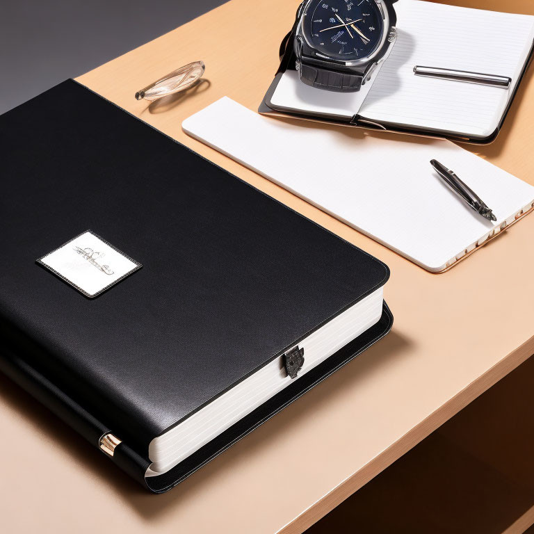 Black Leather Notebook with Silver Emblem, Pen, Wristwatch, and Notepad on Wooden Desk