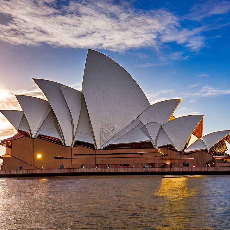 Iconic Sydney Opera House at sunset with sail-like design against blue sky and tranquil harbor reflection