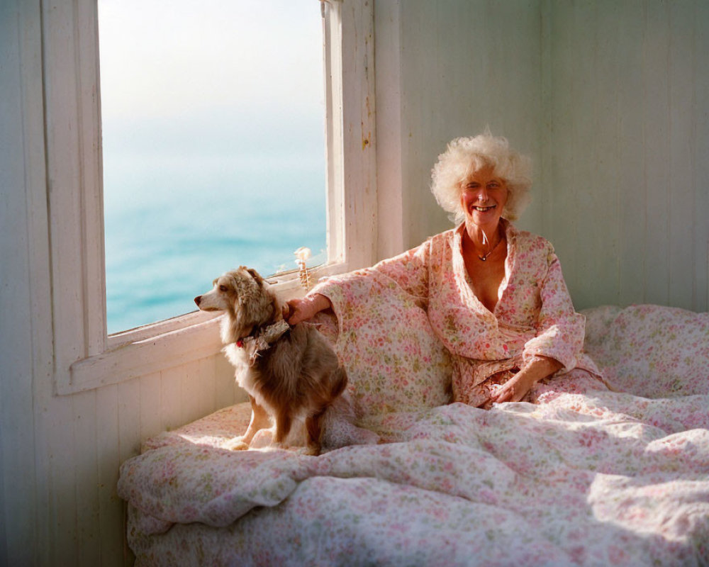 Elderly woman with white curly hair smiles on floral bedspread by ocean view window, petting