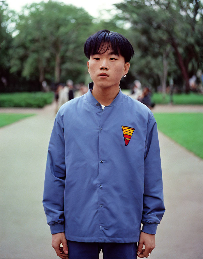 Young person with bowl haircut in blue shirt with emblem, standing in park setting