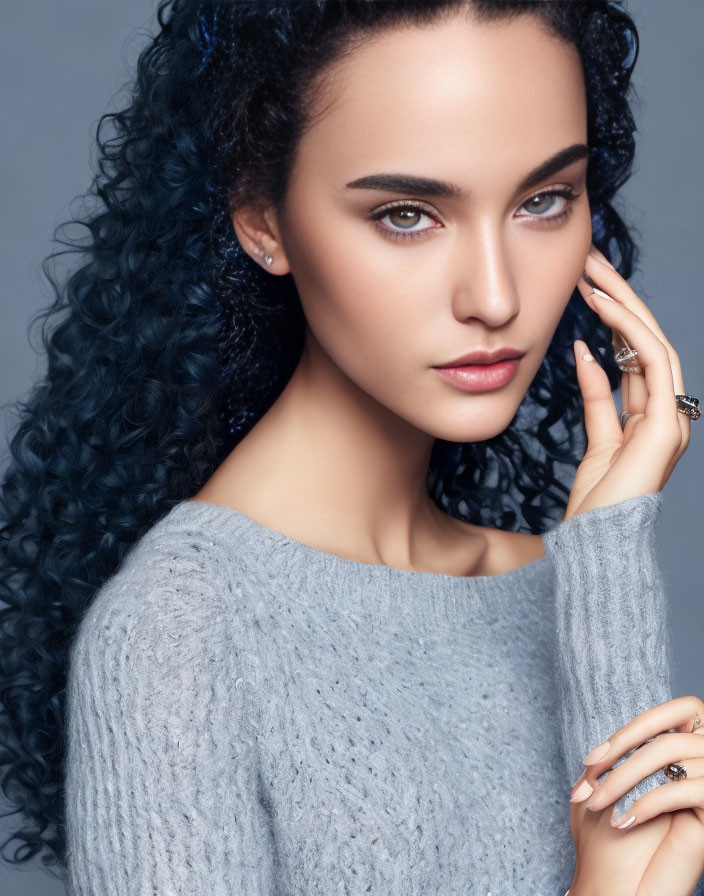 Curly blue-haired woman in grey sweater and rings posing with hand near face