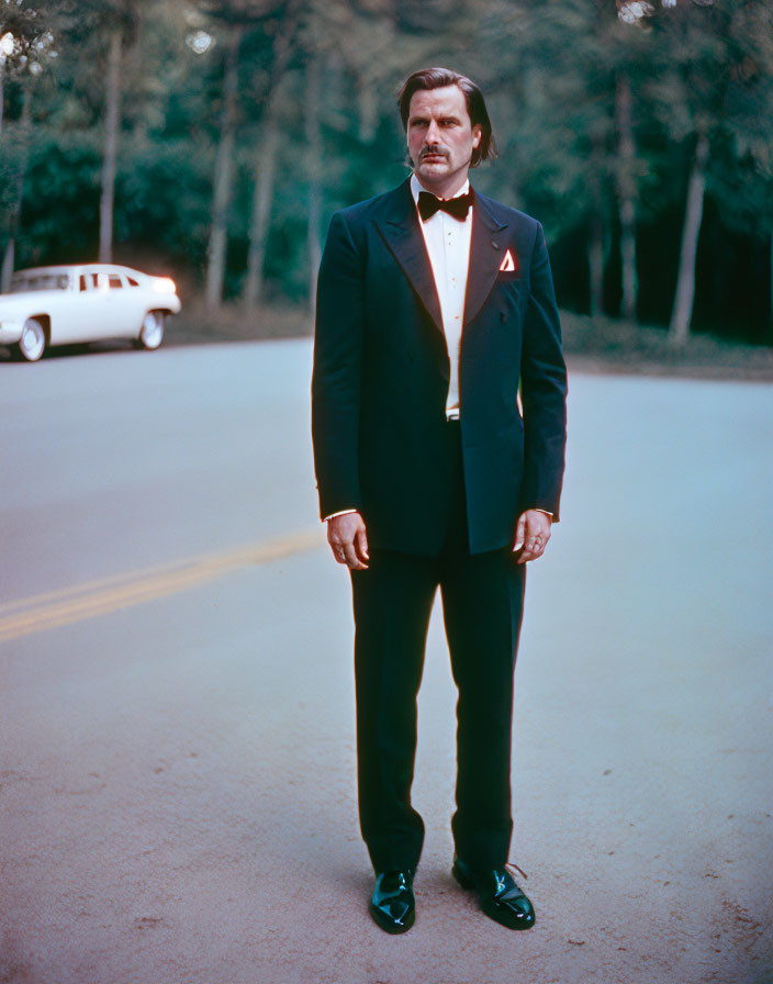 Man in Black Tuxedo with Red Pin Standing by Vintage Car and Trees