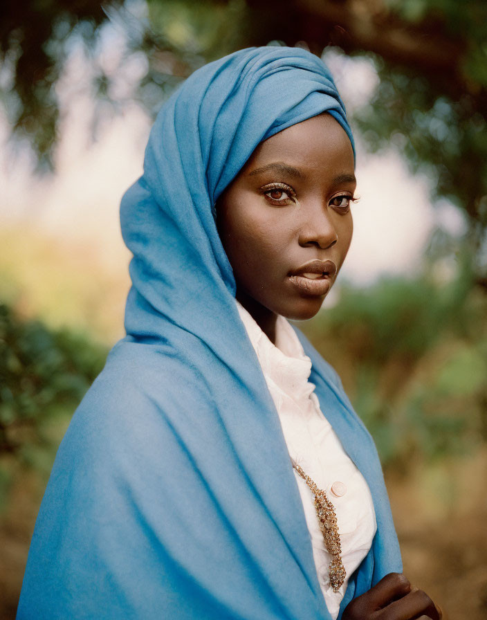 Dark-skinned woman in blue headscarf and white shirt with necklace against blurred nature backdrop